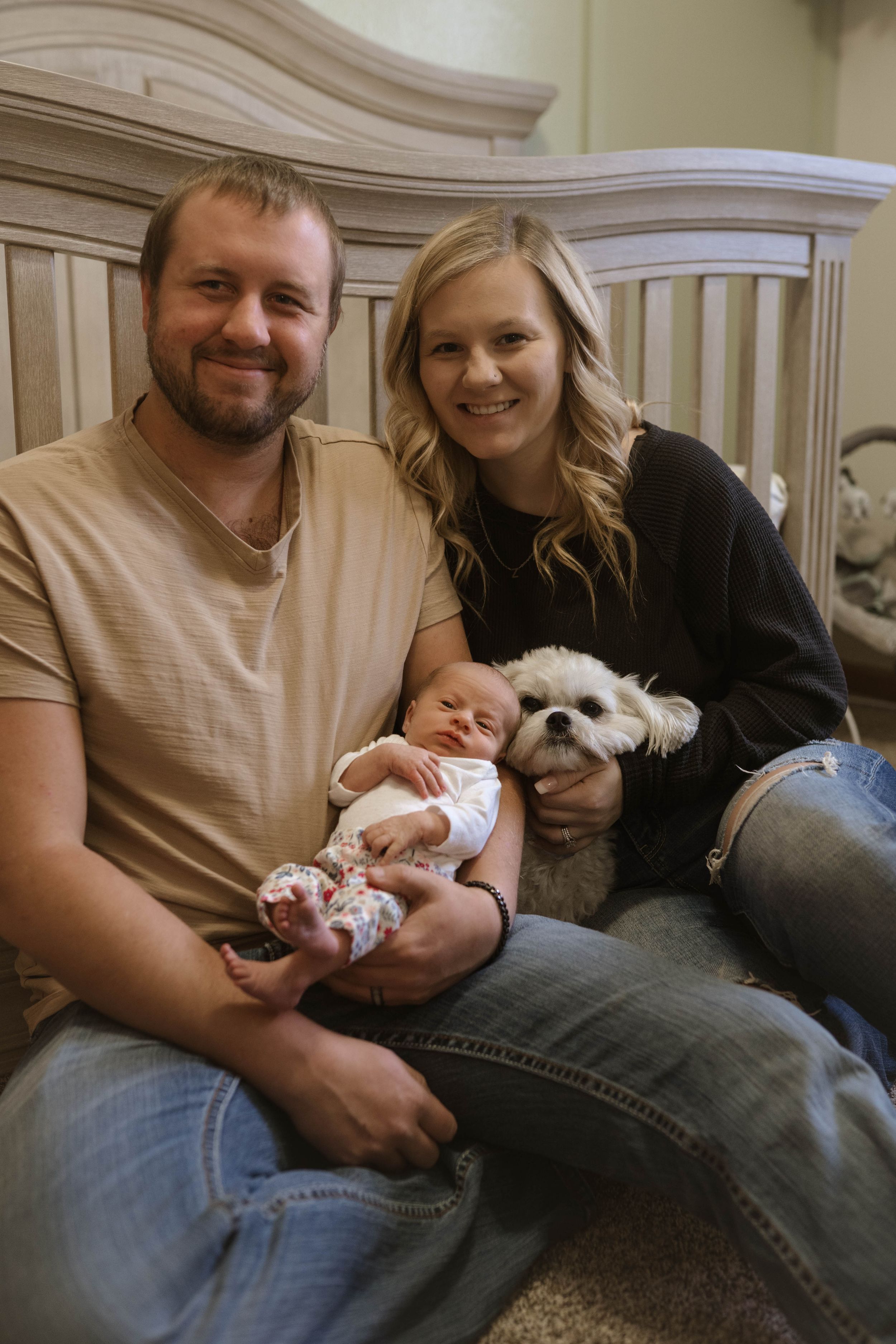 Family portrait with small white dog and newborn baby in nursery setting.
