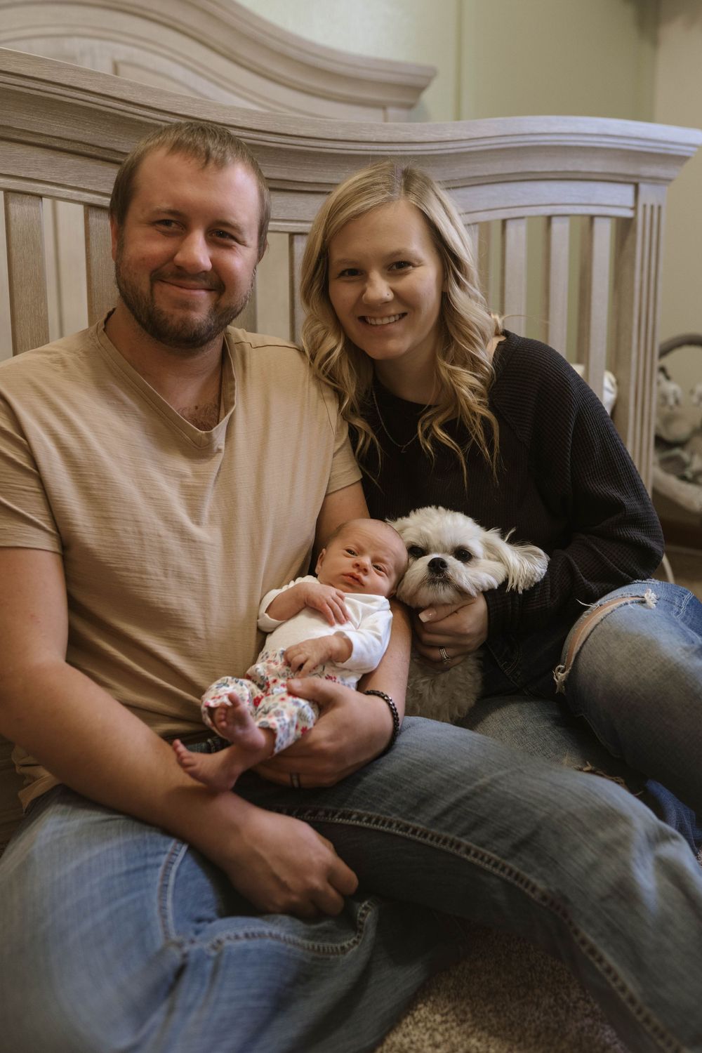 Family portrait with small white dog and newborn baby in nursery setting.