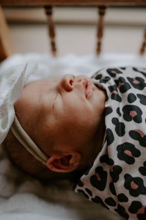 Peaceful newborn resting in a crib wearing a white bow and animal print swaddle.