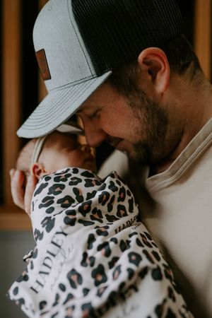 Close up of someone wearing a baseball cap holding a baby wrapped in a leopard print blanket.