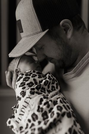 Person in gray cap tenderly holding infant swaddled in animal print fabric.