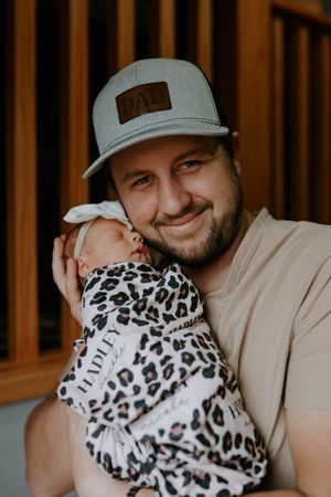 Parent wearing baseball cap sharing intimate moment with newborn in leopard print swaddle.