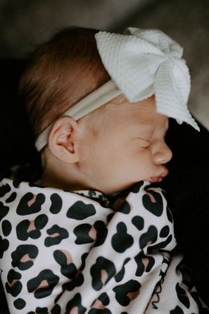 Sleeping newborn wearing white bow headband wrapped in leopard print blanket.