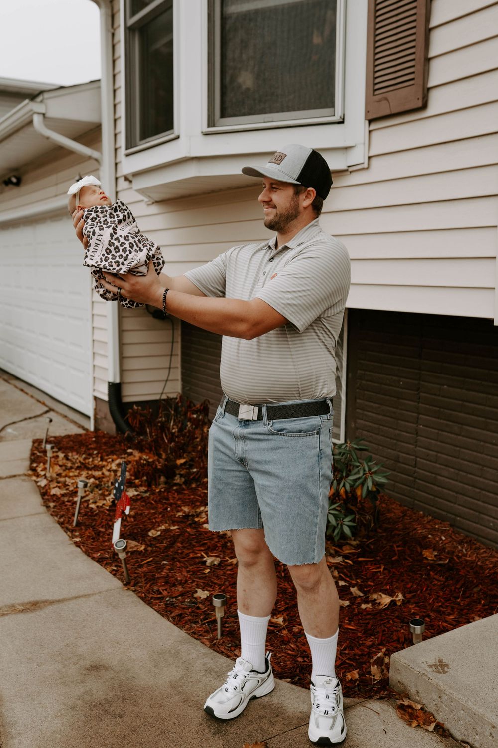 Person in casual attire holding a small item near a white house with red mulch landscaping.