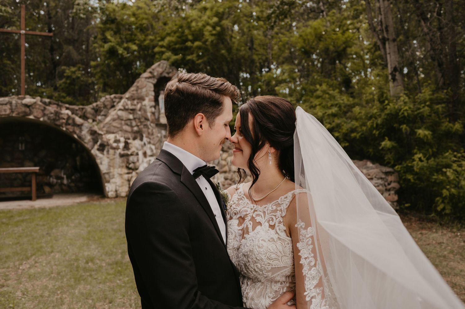 A romantic wedding couple shares an intimate moment in front of a stone wall surrounded by lush greenery.