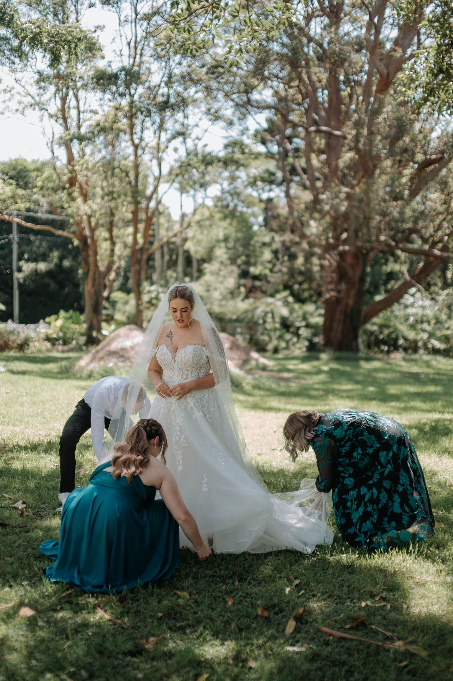 Wedding party in teal bridesmaid dresses relaxing on grassy lawn under eucalyptus trees on sunny afternoon.