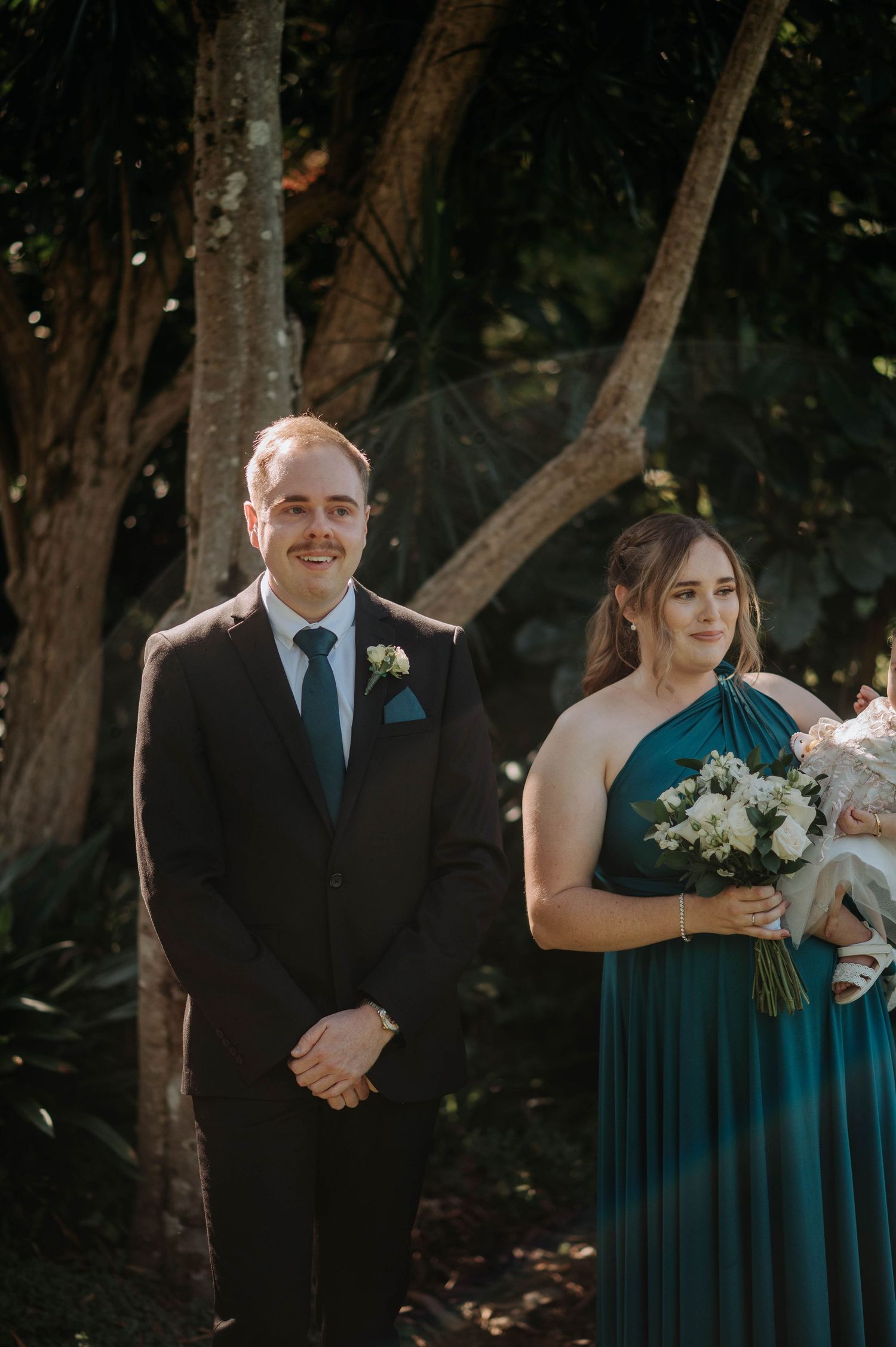 Wedding couple and bridesmaid in teal dress pose together in a wooded outdoor setting during golden hour.