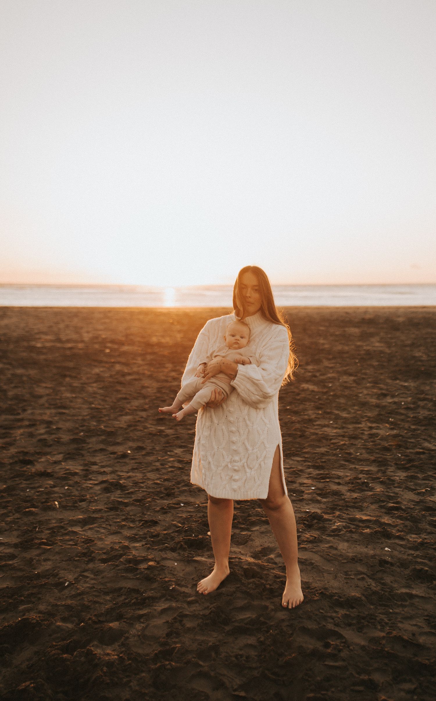 Smiling Nami Julia Hampe in cream knit dress holding baby on dark sandy beach at golden sunset in New Zealand.