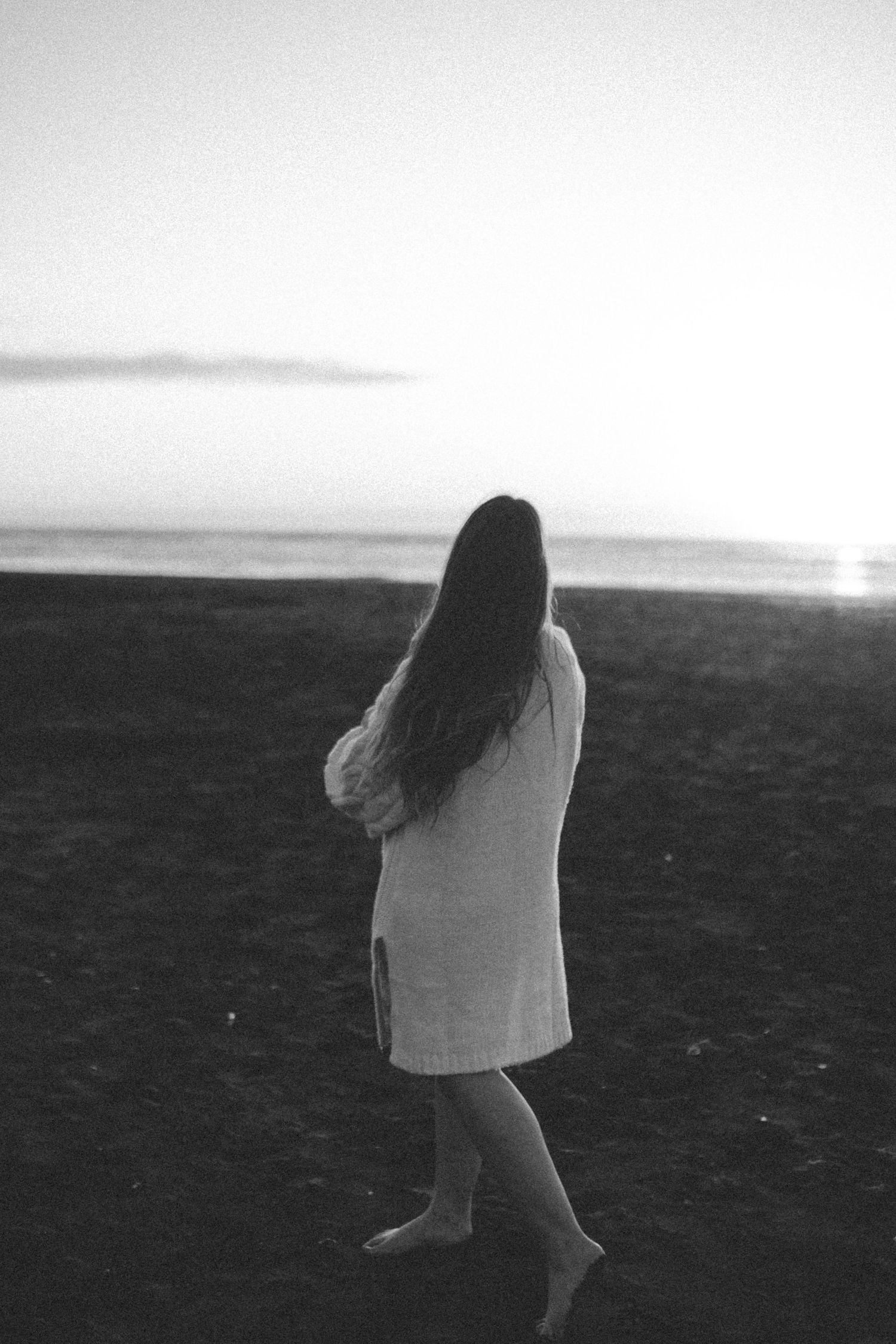 Black and white image of Nami Julia Hampe in white dress walking alone on beach at sunset in New Zealand.