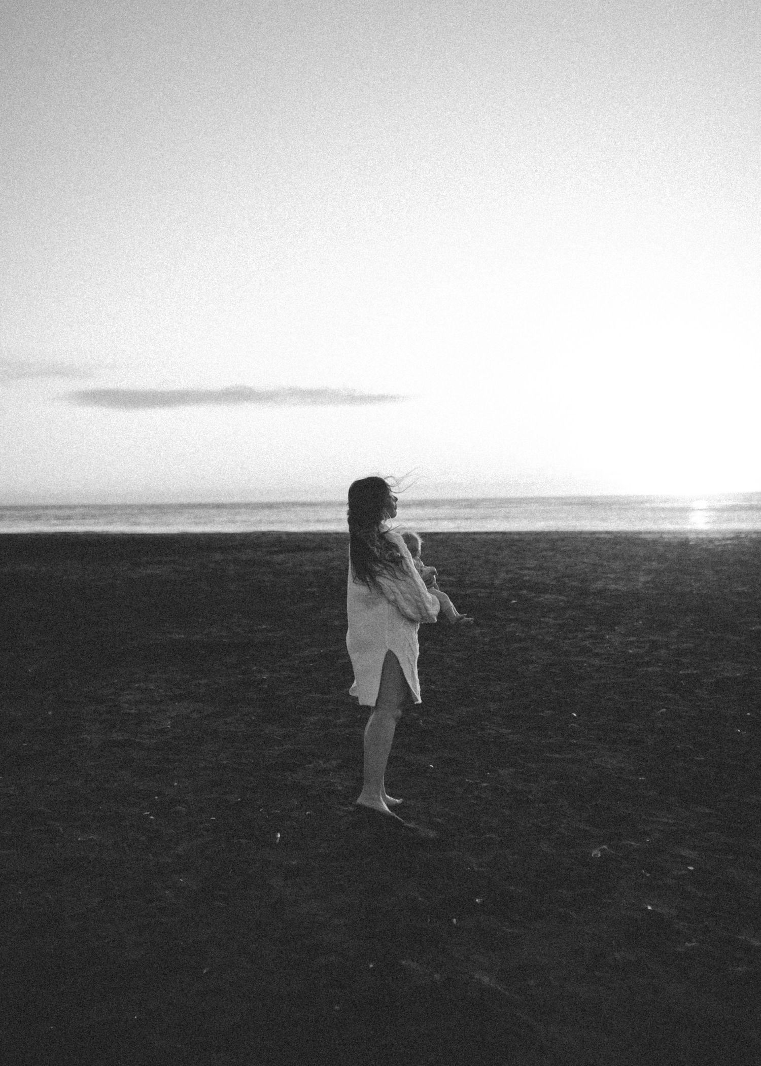 Black and white image of Nami Julia holding baby on open beach during peaceful sunset  in New Zealand.