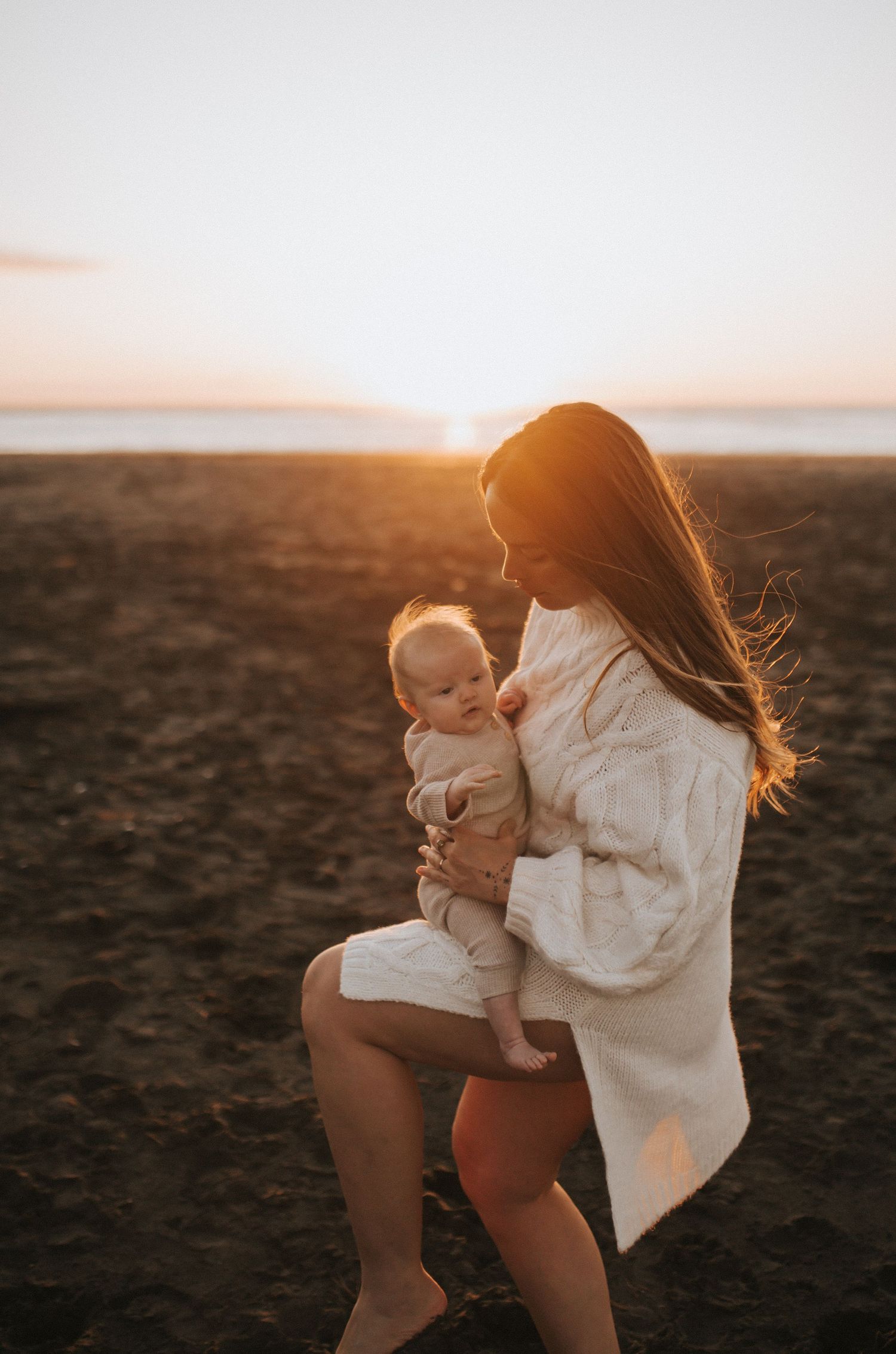 Nami Julia Hampe in cream knit cardigan cradling baby on dark sandy beach during golden hour sunset in New Zealand.