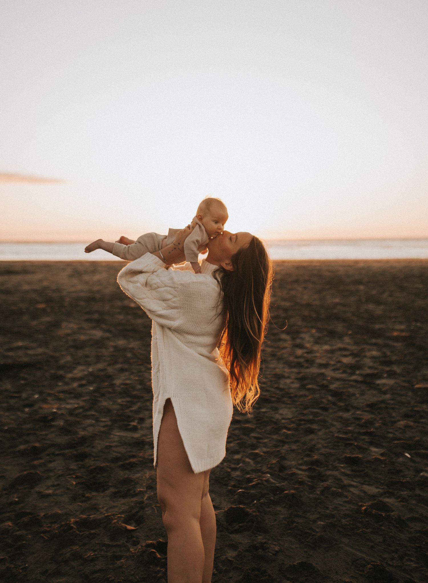Nami Julia Hampe joyfully lifting baby overhead on sandy beach during vibrant golden hour sunset in New Zealand.