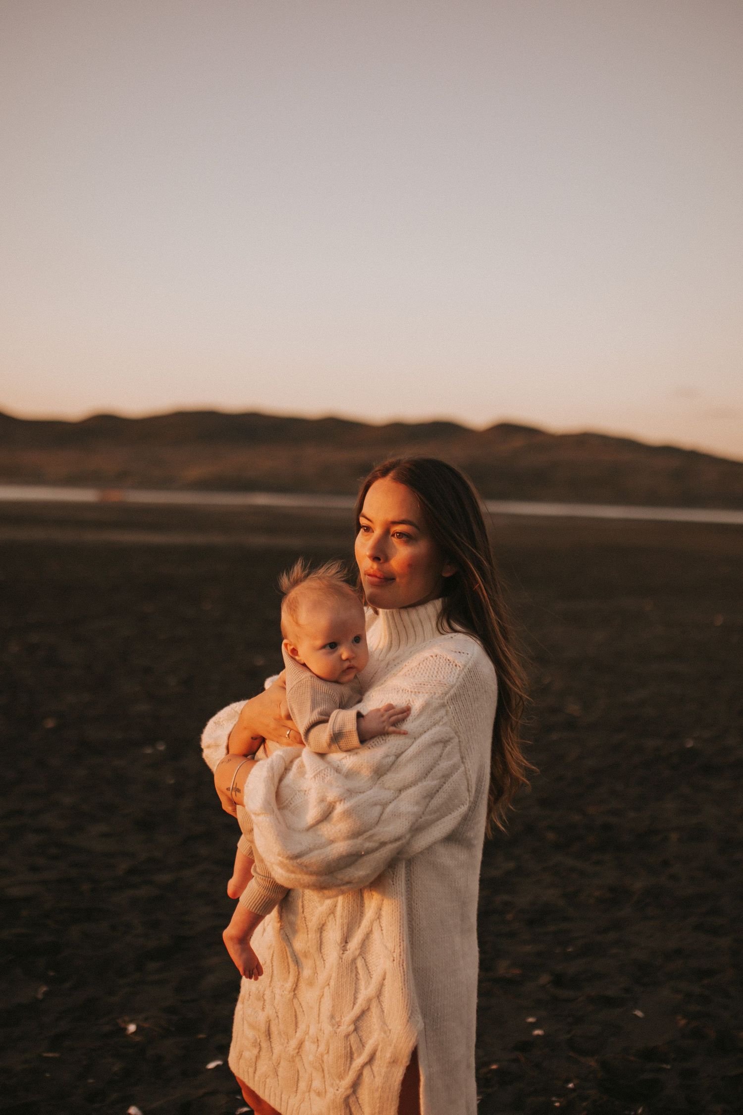 Nami Julia Hampe in a white cable-knit sweater holds her baby on a beach at golden hour sunset  in New Zealand.