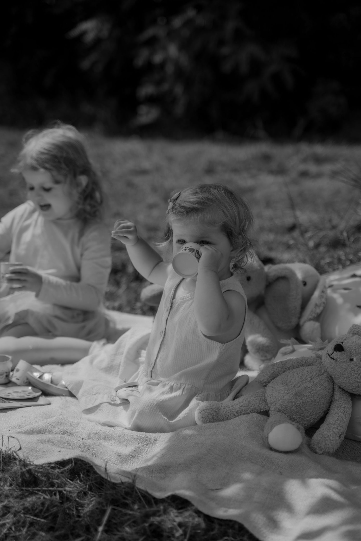 A vintage black and white photograph of children having a picnic outdoors with their teddy bear on a blanket.