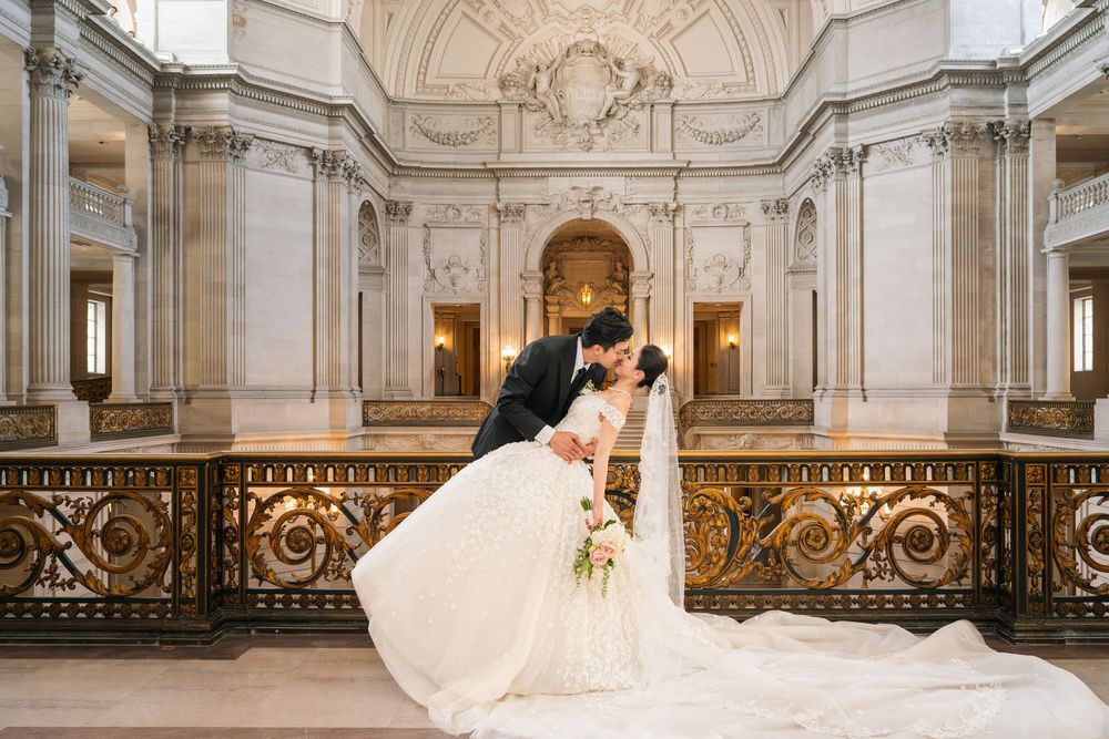 Elegant wedding couple dances in grand marble ballroom with ornate architecture and sweeping staircase.