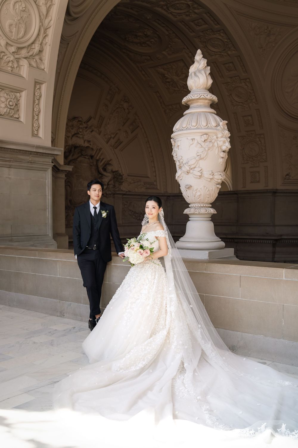 Elegant wedding couple poses by ornate stone urn in grand architectural setting with flowing white gown.