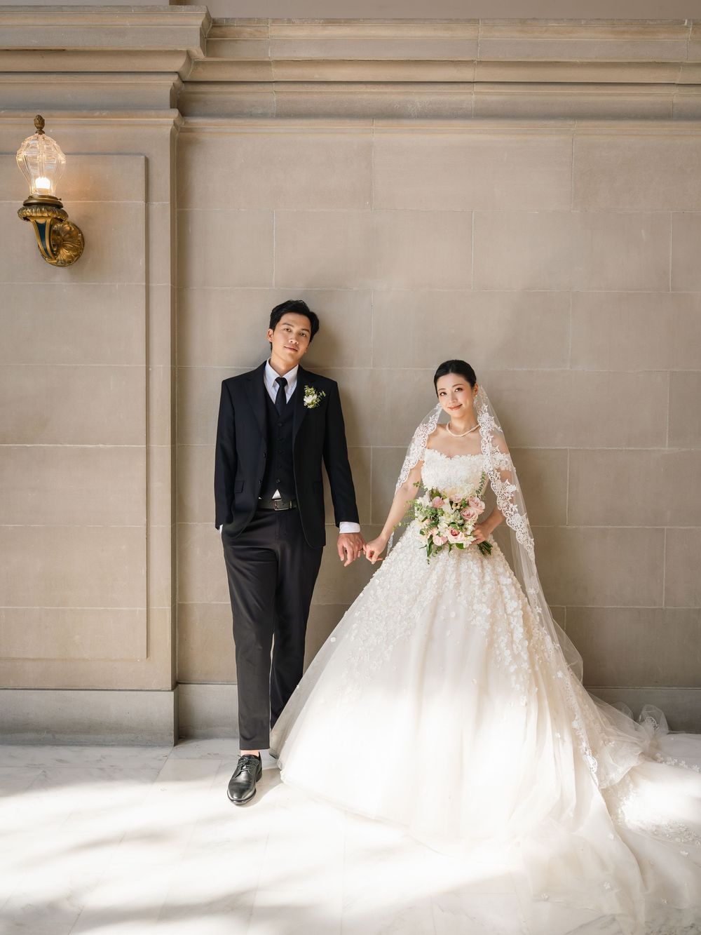 A romantic wedding photo series showing a couple in formal attire against a light stone wall with elegant lighting.