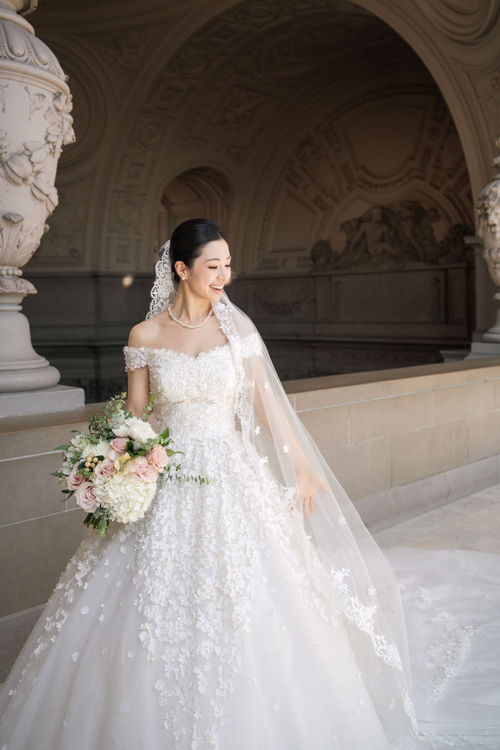 A bride in a white lace wedding gown with floral appliques poses elegantly in a grand arched hallway.