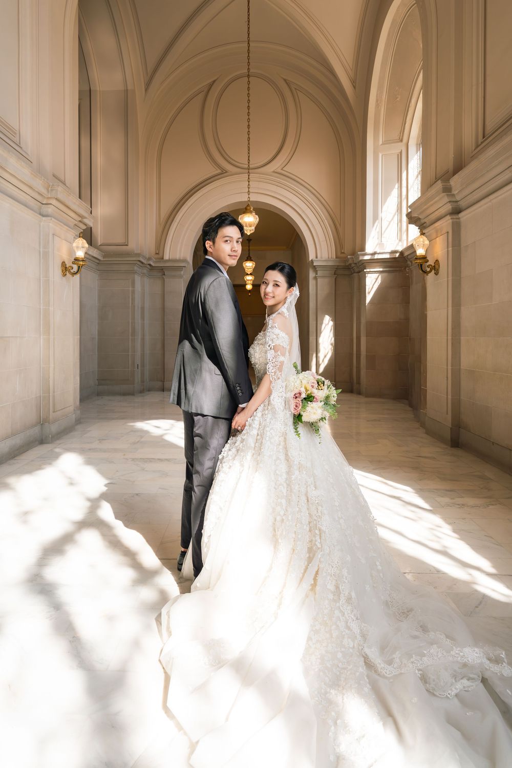 Romantic wedding portrait in sunlit marble hallway with arched ceilings and classic architecture.