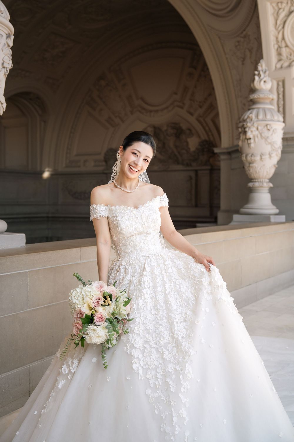 A bride in a luxurious white ballgown poses elegantly in an ornate architectural setting with decorative columns.