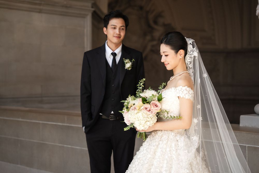 Elegant wedding couple poses in formal attire against stone columns in a classic architectural setting.