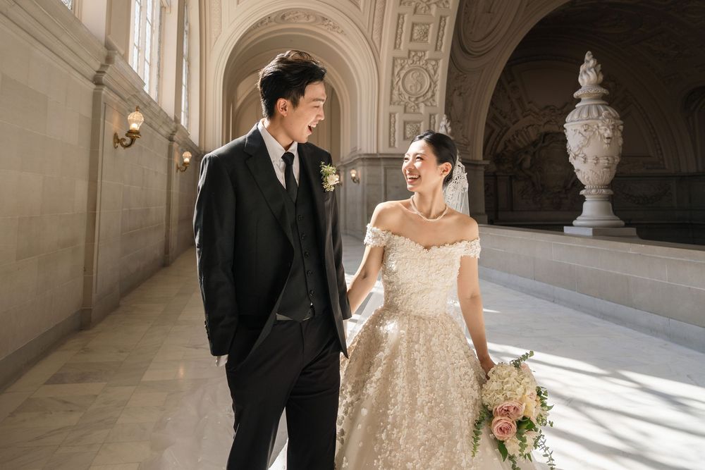 A couple in wedding attire walks together through an elegant corridor with arched architecture and natural lighting.