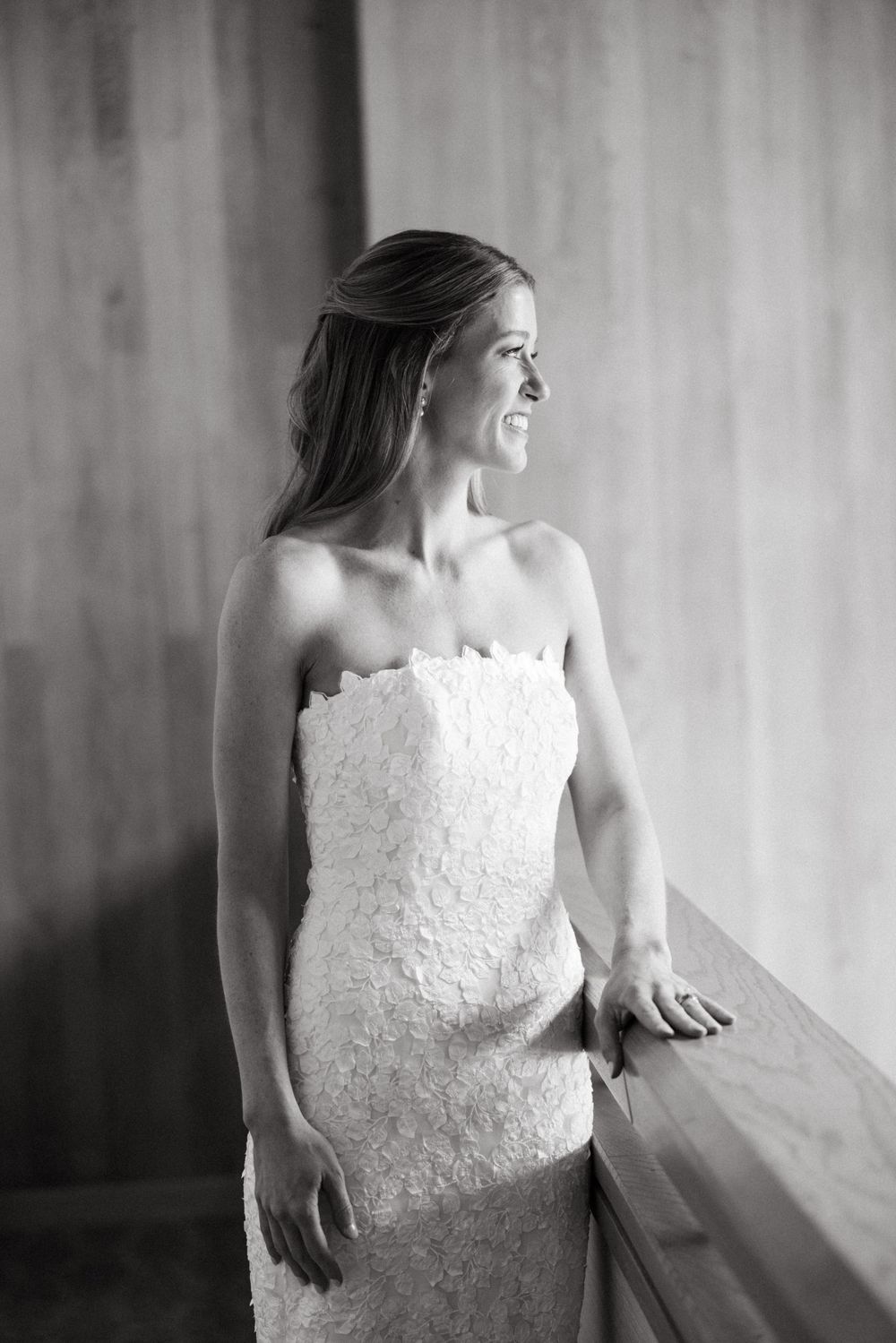 Elegant bride in a strapless lace wedding gown gazes out a window in black and white portrait.