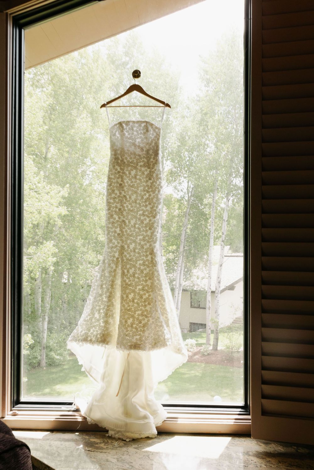 White lace wedding dress hangs elegantly by a window with natural light streaming in, creating a romantic silhouette.