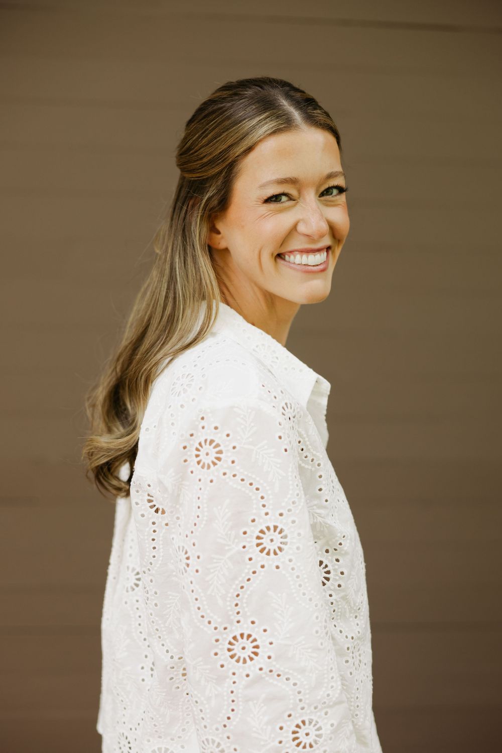 Woman wearing a white eyelet lace top poses with a bright smile against a neutral background.