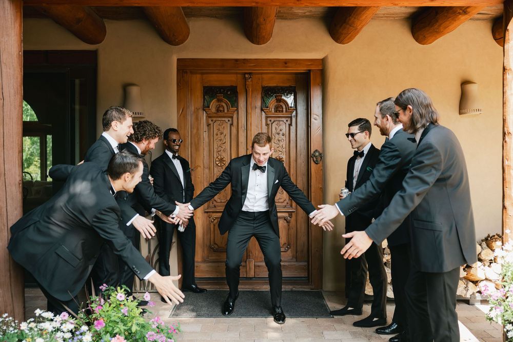 A sequence of groomsmen perform a coordinated jumping pose in formal attire on a porch.