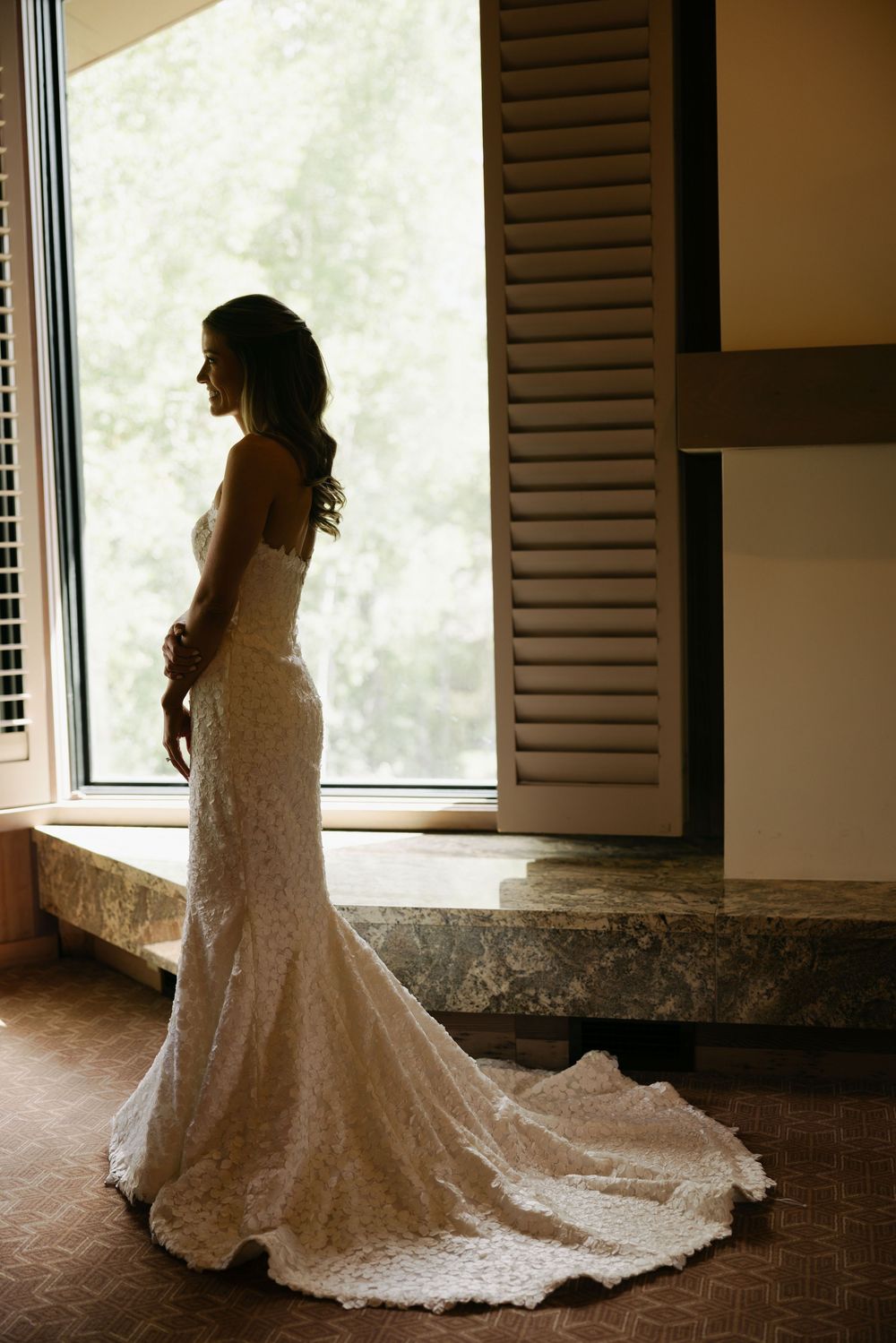 A stunning lace wedding dress with a flowing train is silhouetted against a bright window in an elegant indoor setting.