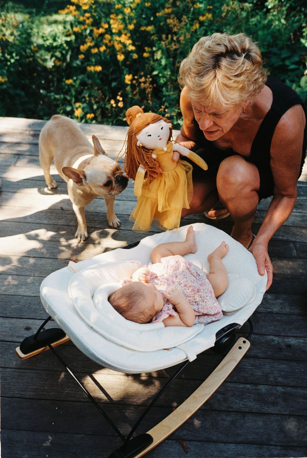 Grandmother rocks her grand daughter outside on the porch with a doll in hand as their French bulldog tries to grab the doll at a party