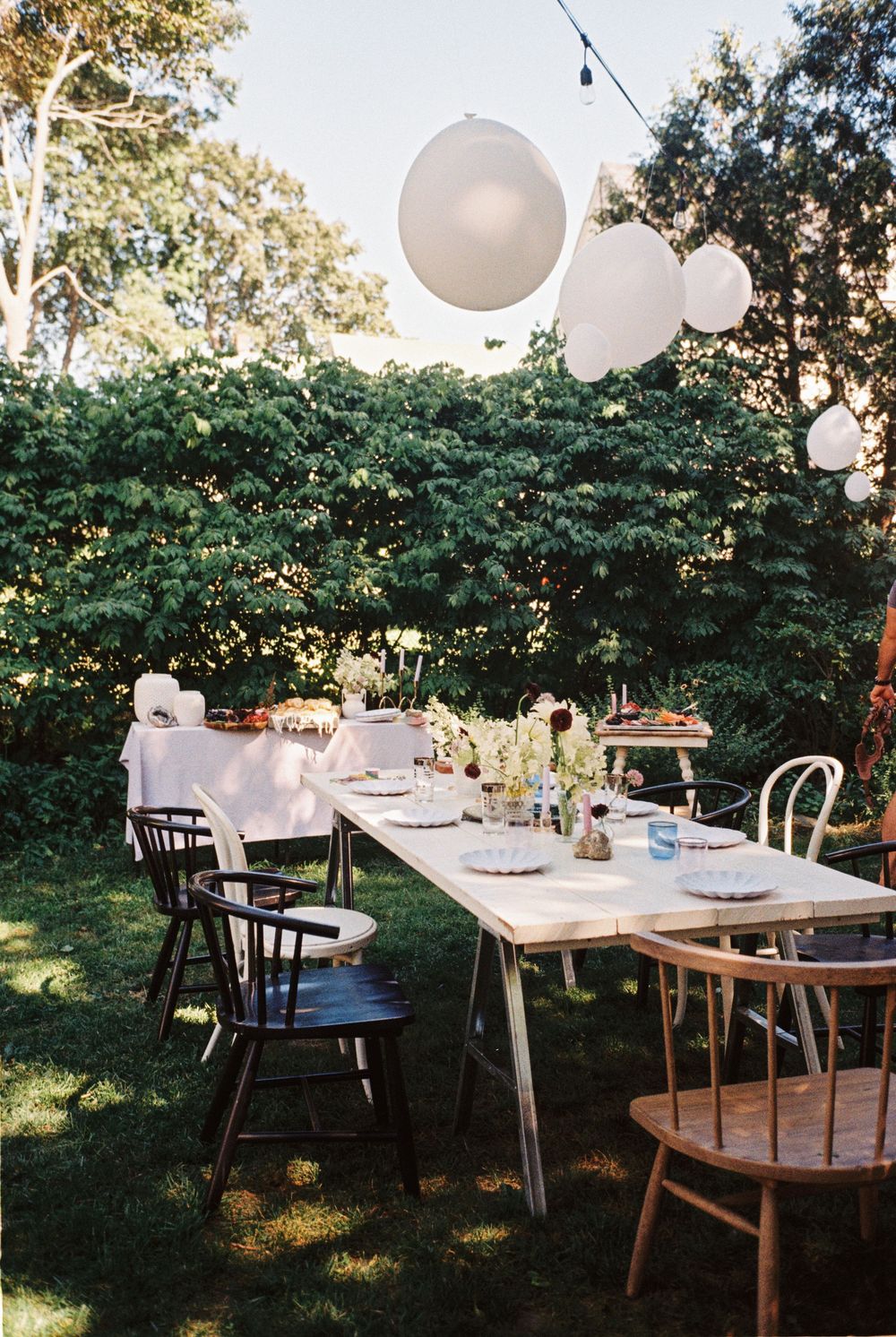 A decorated table with midcentury modern mismatched chairs and lilac pastel tableware with a string of balloons hanging at a baby shower.