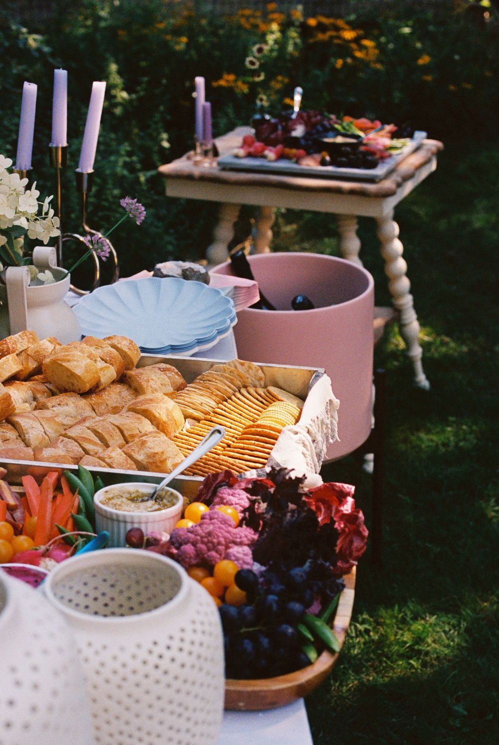 Colorful charcuterie of vegetables and fresh baked breads next to crudités and pastel lilac plates and pink buckets of champagne at party.