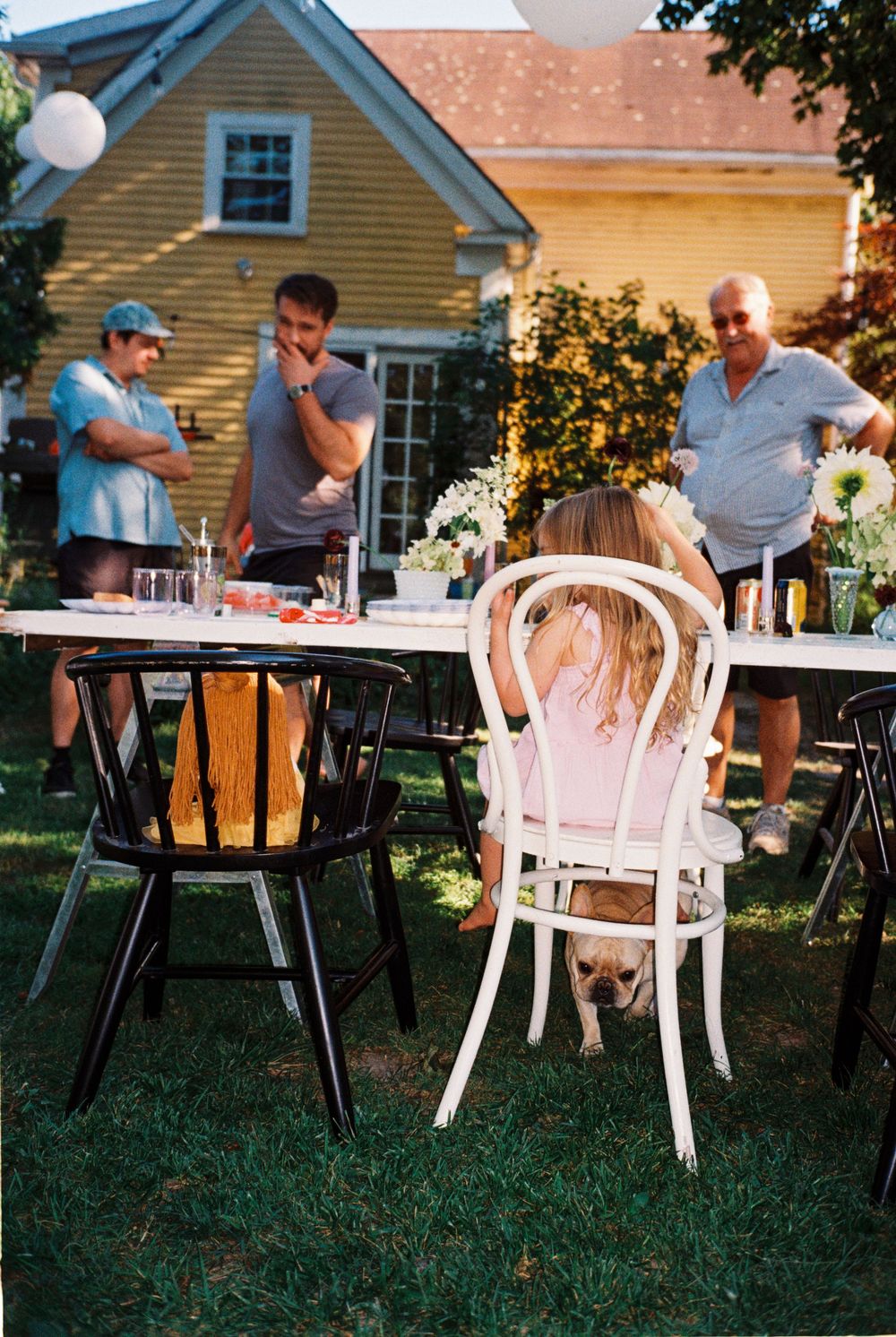 Little girl sits in a chair with her doll seated in the chair next to her while people chat & smile during a sprinkle baby shower in August.