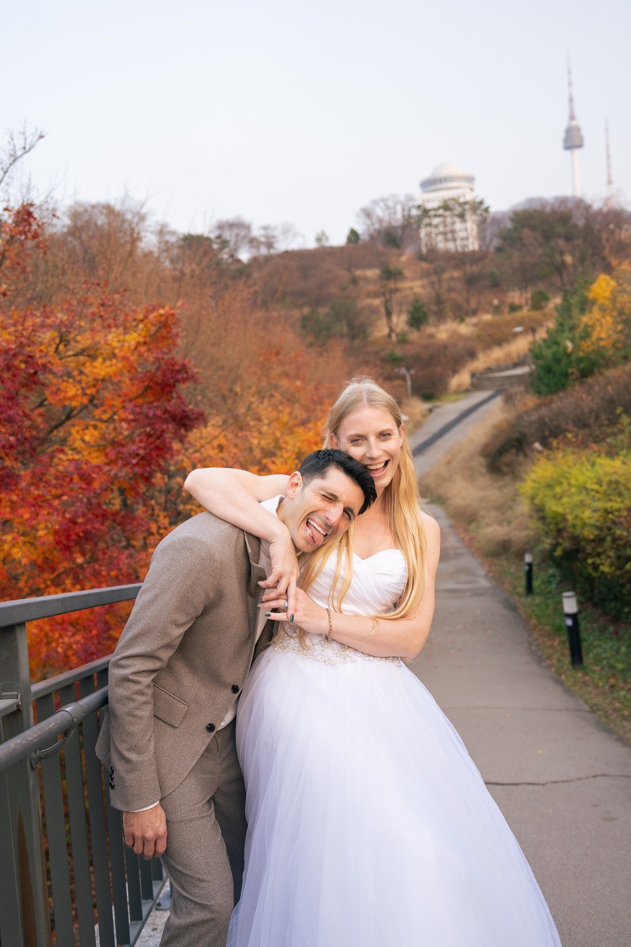 A couple in wedding attire poses playfully on a path surrounded by vibrant autumn foliage and fall colors.
