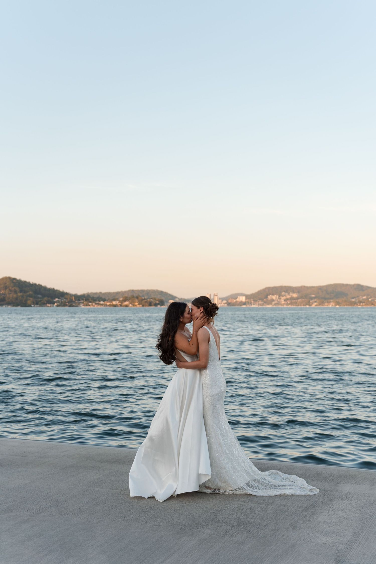 Two people in white dresses share an intimate moment by the ocean at sunset with mountains in the background.