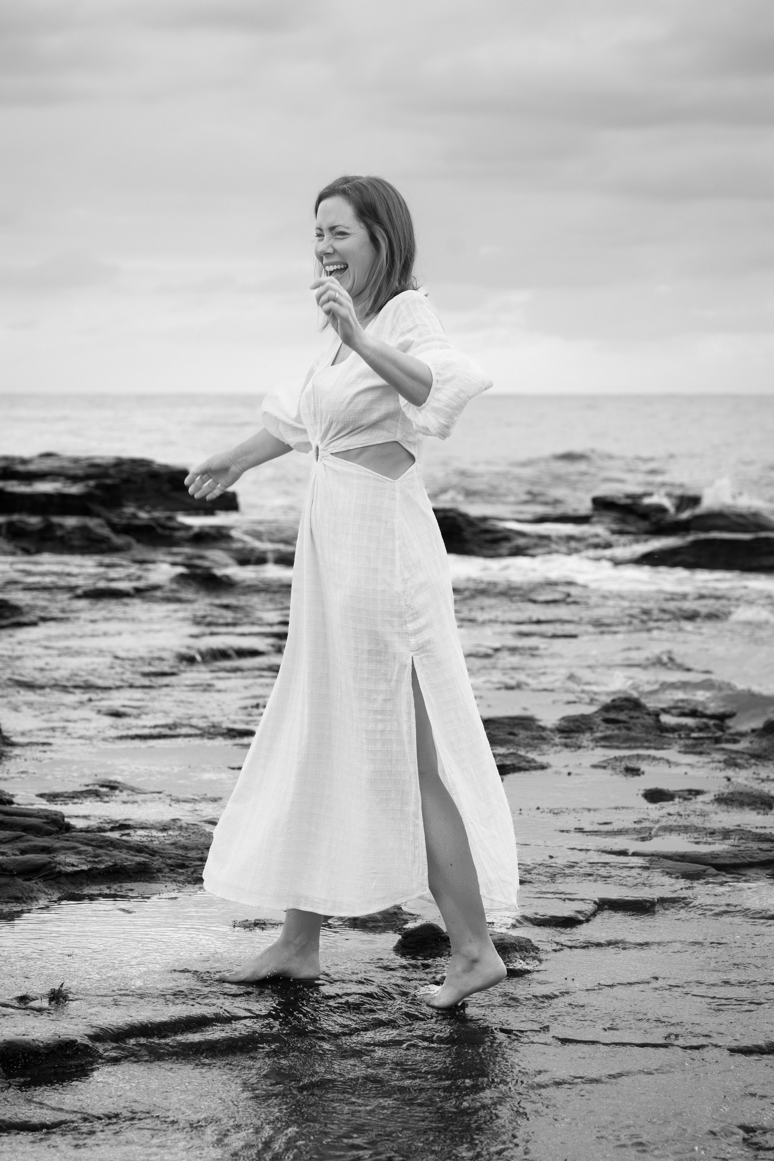 Black and white photo series of someone in a flowing white dress dancing and twirling on rocky beach shoreline.