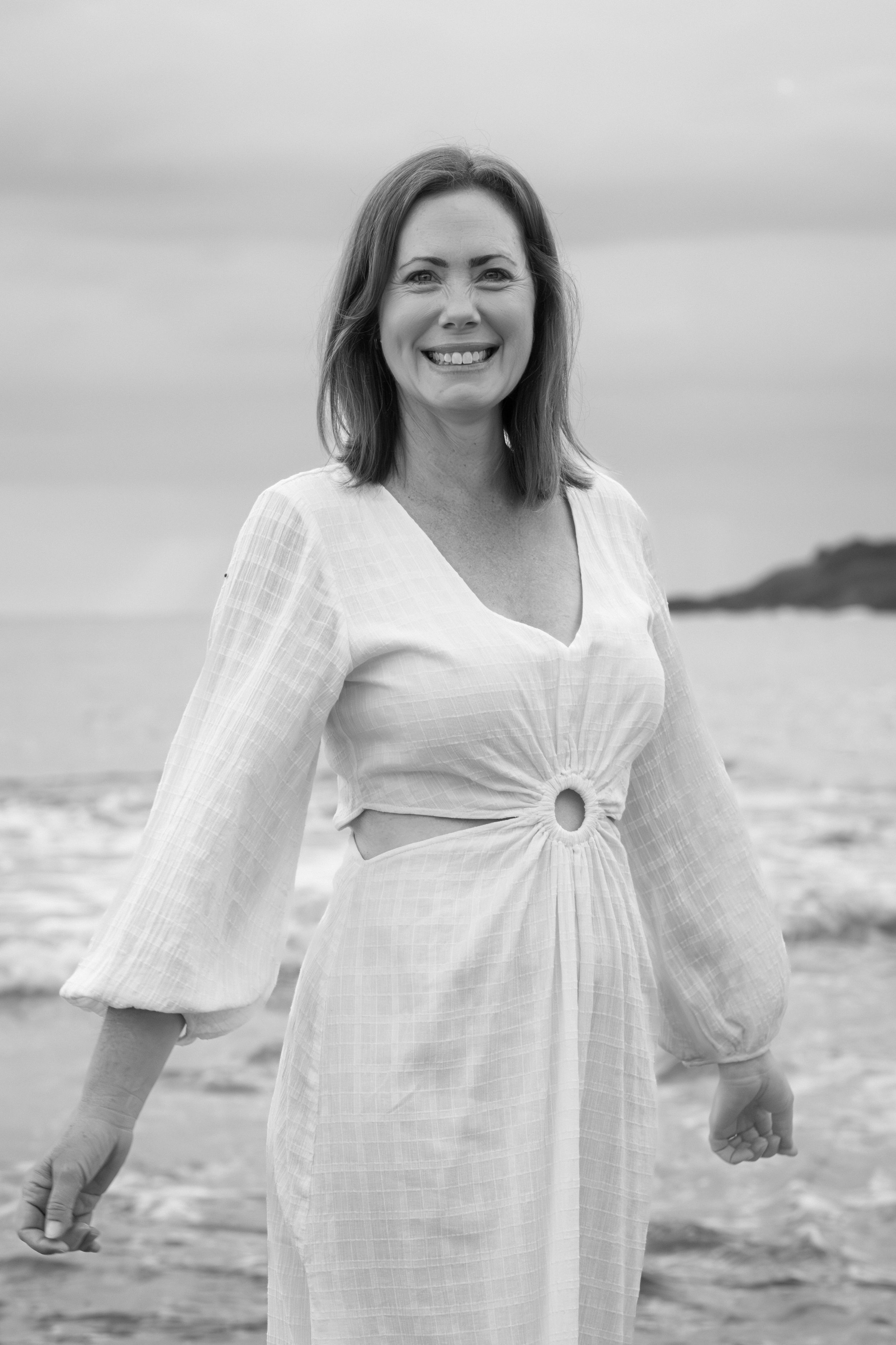 A person in a white dress smiles and walks along a beach shore in black and white photography.