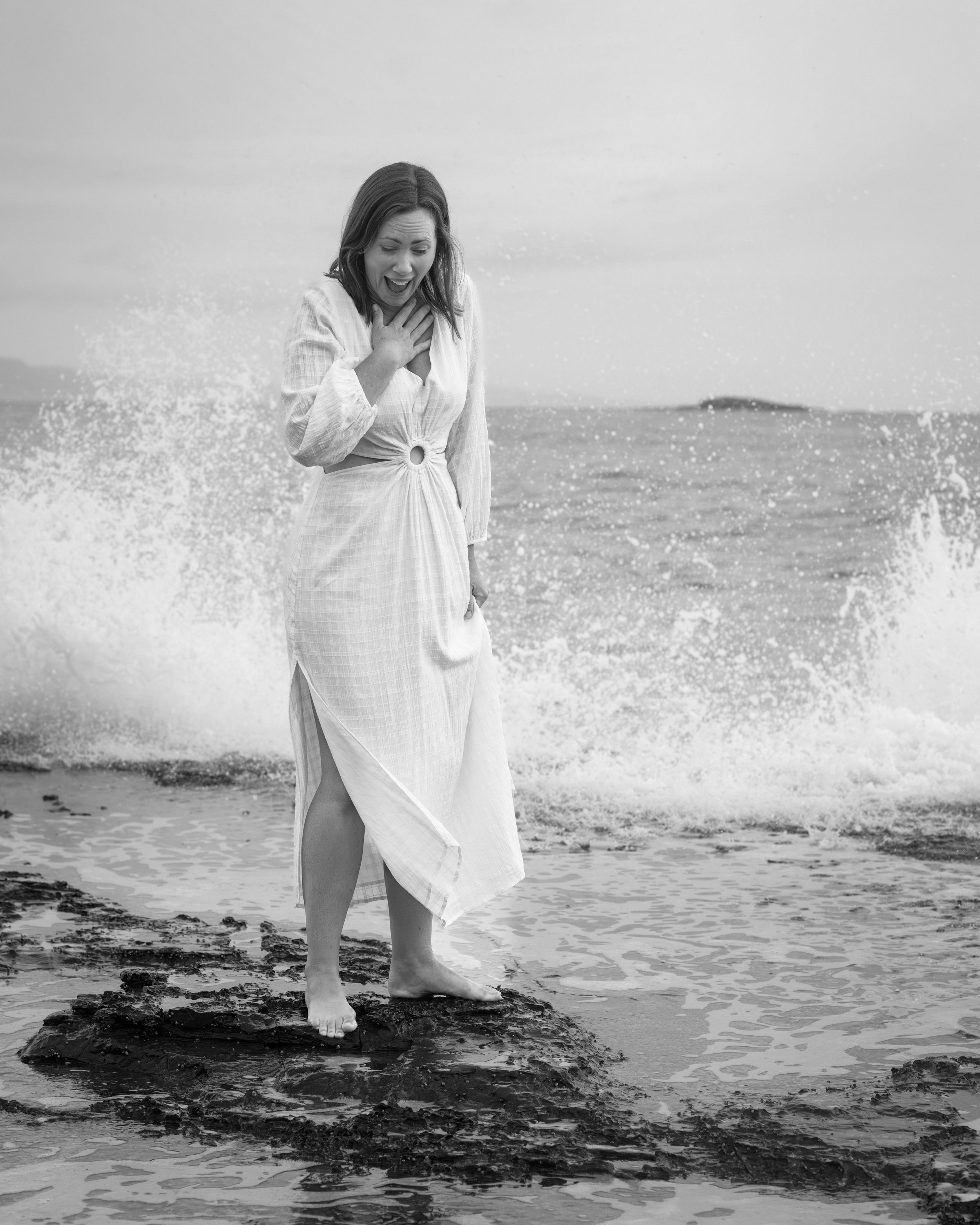 A figure in a flowing white dress stands on dark rocks as ocean waves crash and spray around them in black and white.