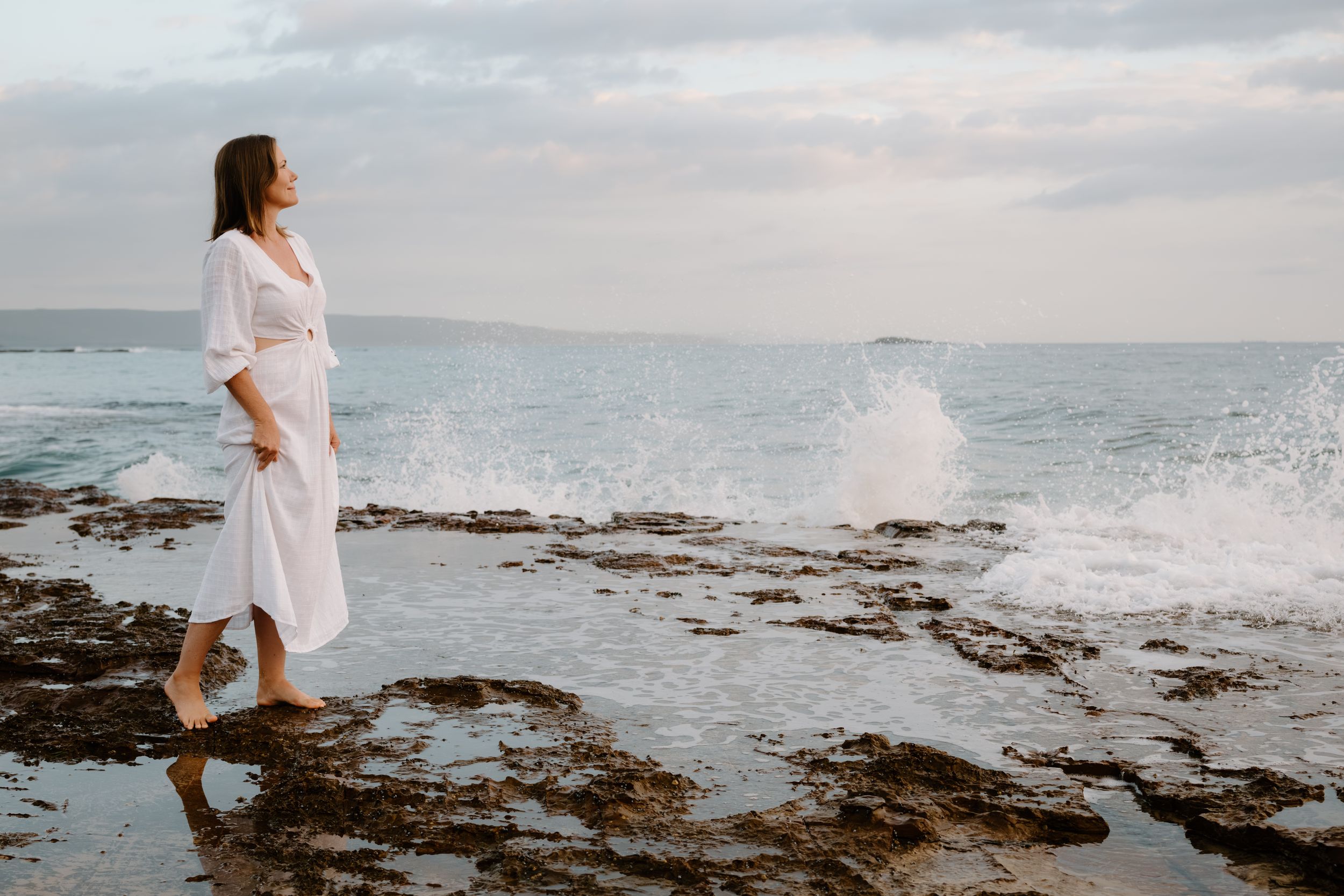 A person in a white dress walks along a rocky beach coastline as waves crash against the shore at sunset.