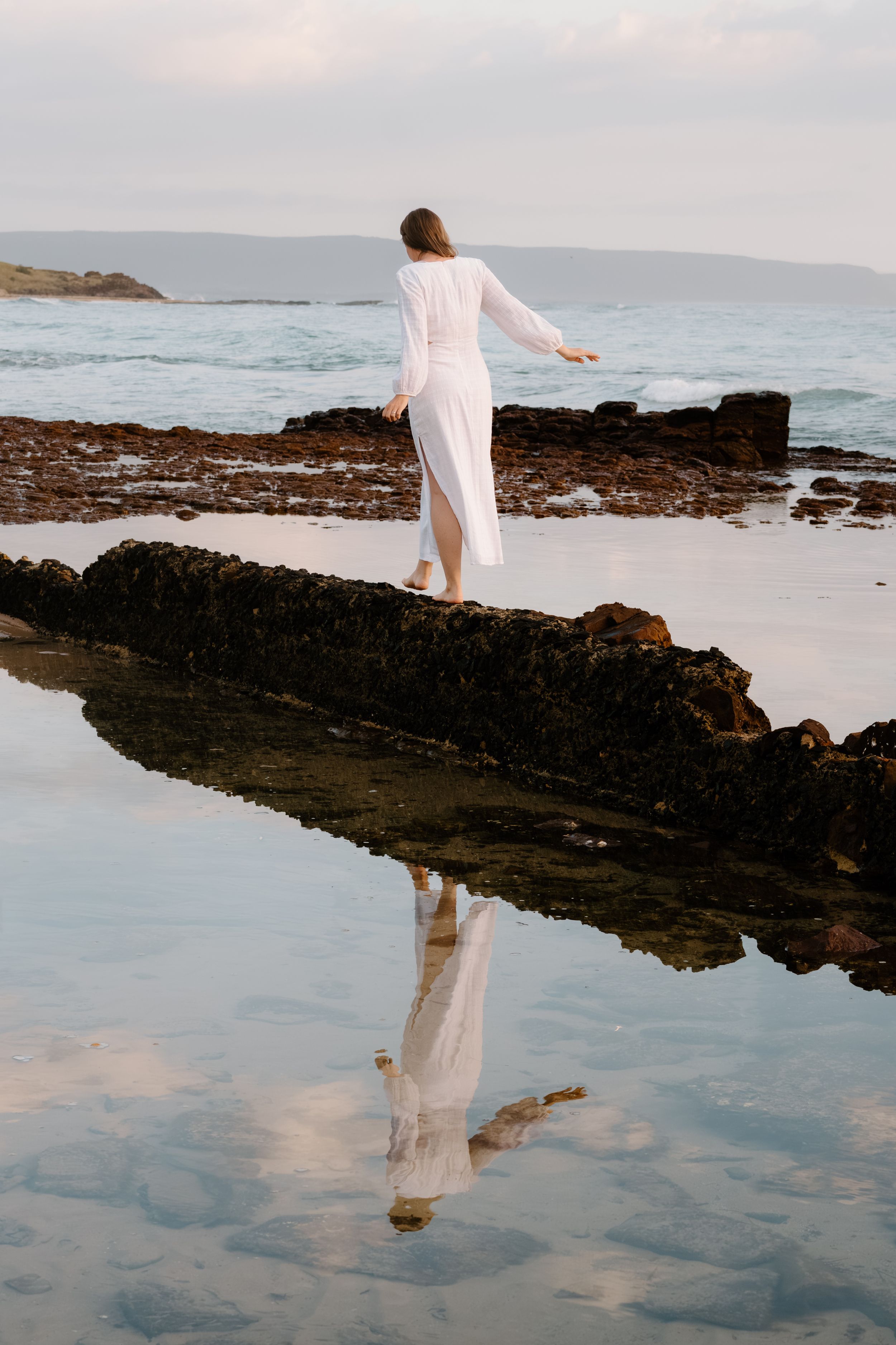 A figure in a flowing white dress stands gracefully on rocky coastal tide pools at sunset, creating a dreamy reflection.