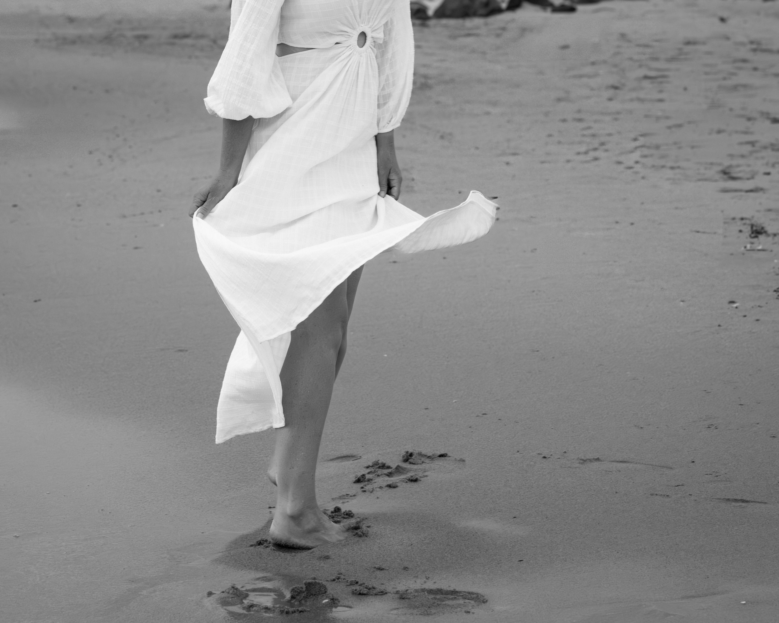 A person in a flowing white dress walks barefoot along a sandy beach leaving footprints in black and white.