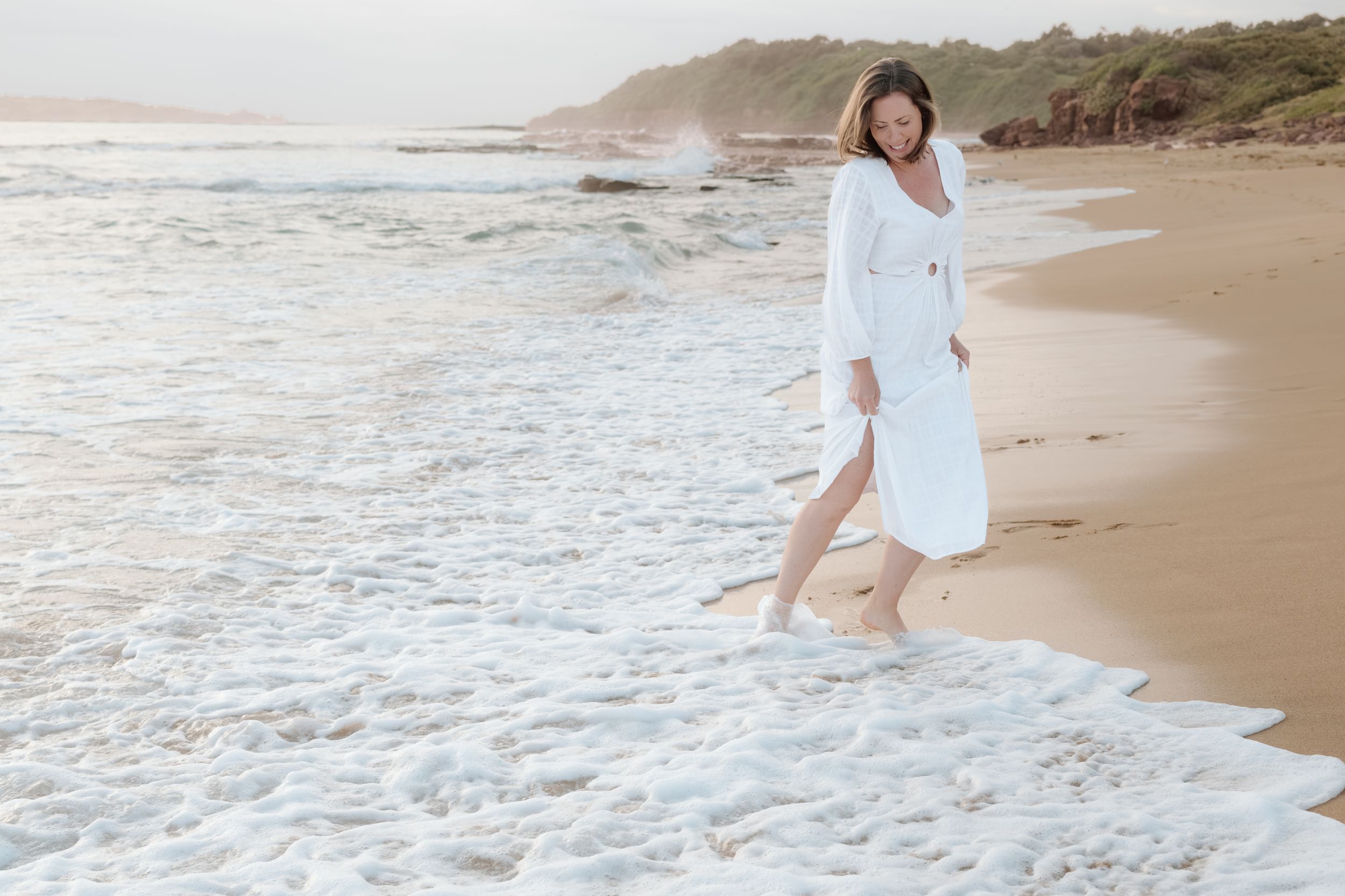 A person in a flowing white dress walks along a serene beach shoreline at sunset, creating a dreamy coastal scene.