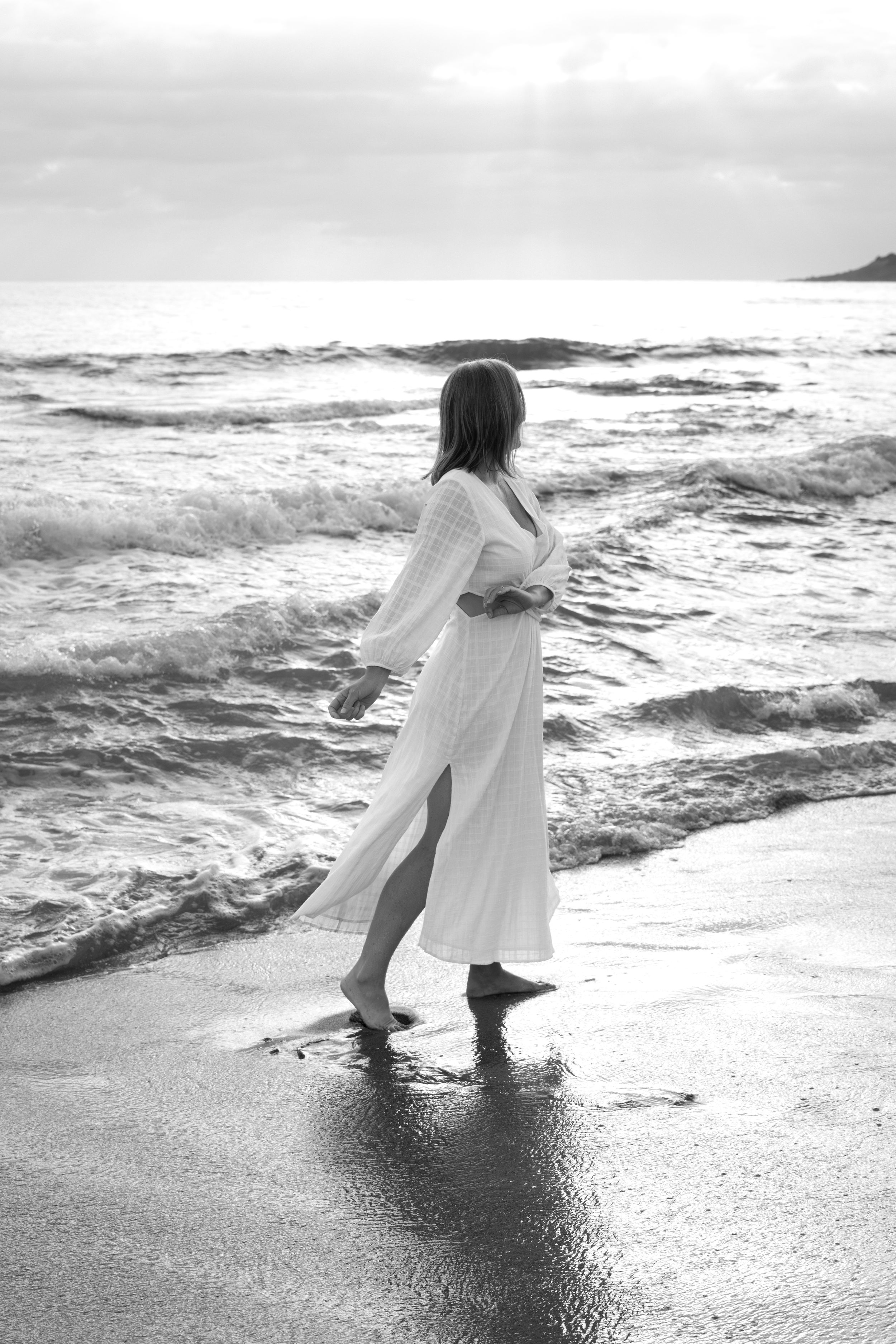 A person in a flowing white dress dances along the beach shoreline as waves crash in black and white photography.
