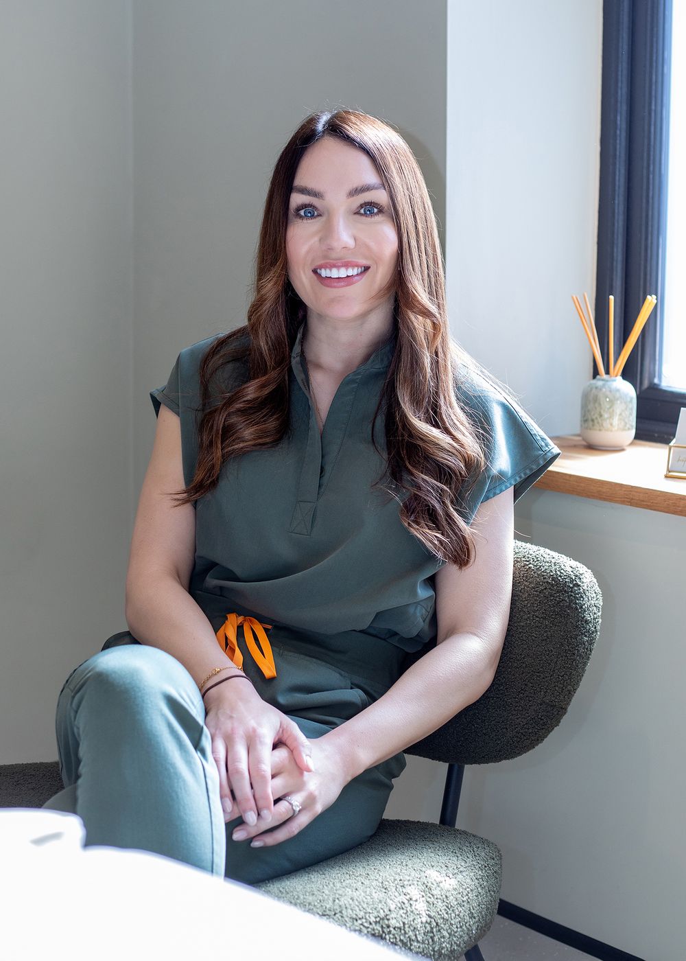 Professional seated in a dark green outfit on a textured gray armchair in a bright modern office setting.