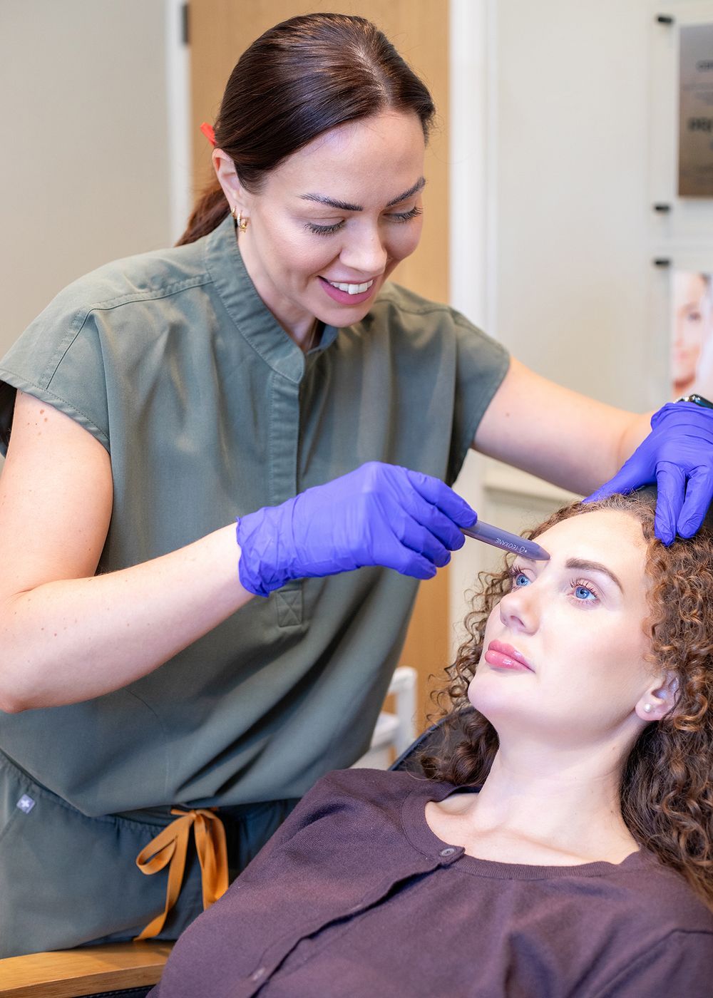 Medical professional in olive scrubs performs cosmetic treatment on patient lying on treatment chair.