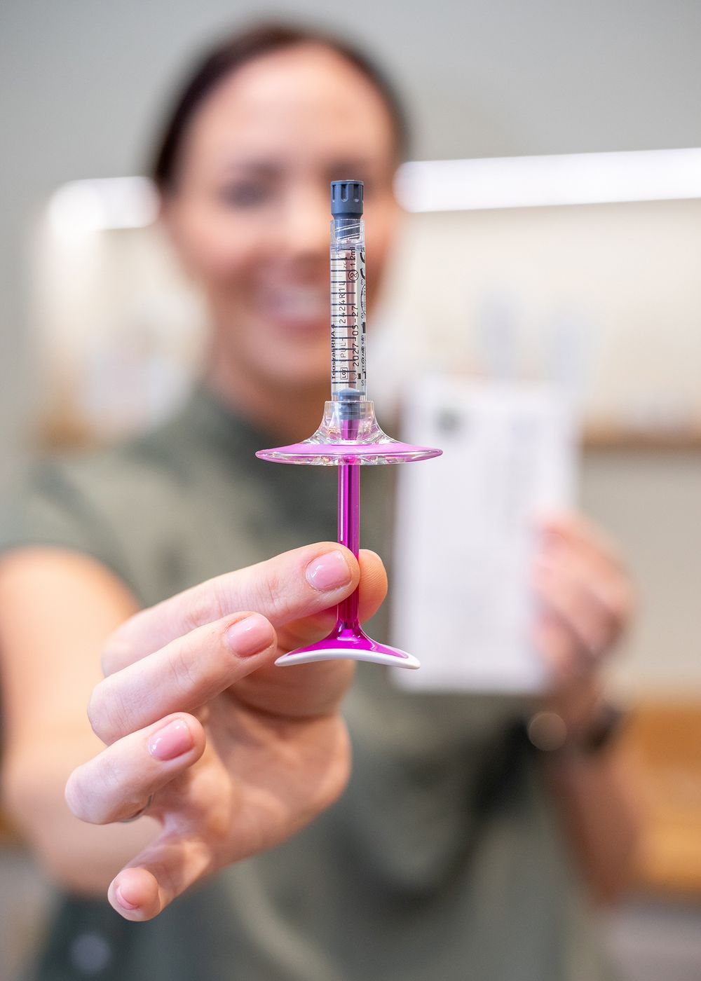 Hand holding a pink martini glass with a syringe-style dropper containing clear liquid against a blurred background.