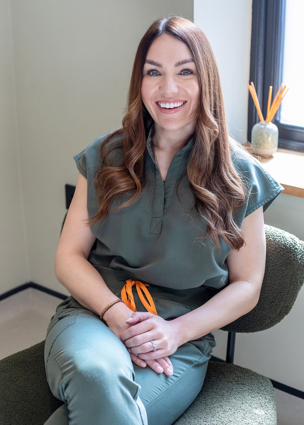 A healthcare professional in sage green scrubs sits smiling in a chair while holding orange scissors.
