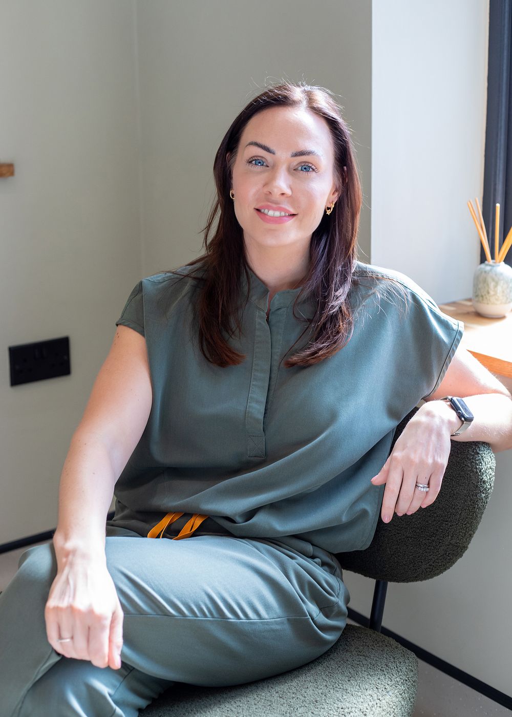 Person in a sage green outfit reclines on a dark green velvet couch in a modern minimalist interior setting.